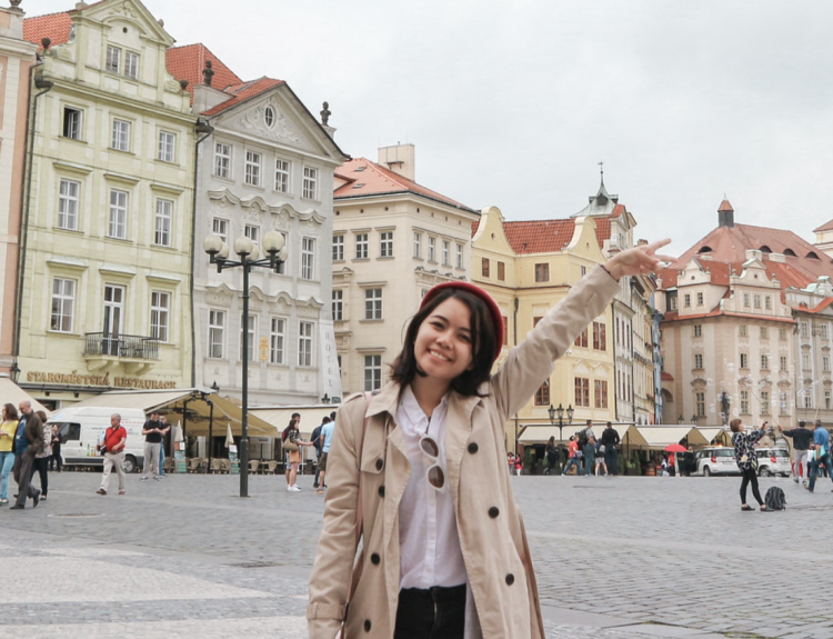 A girl with a peace sign, with beautiful buildings in the background. Taken in Prague.