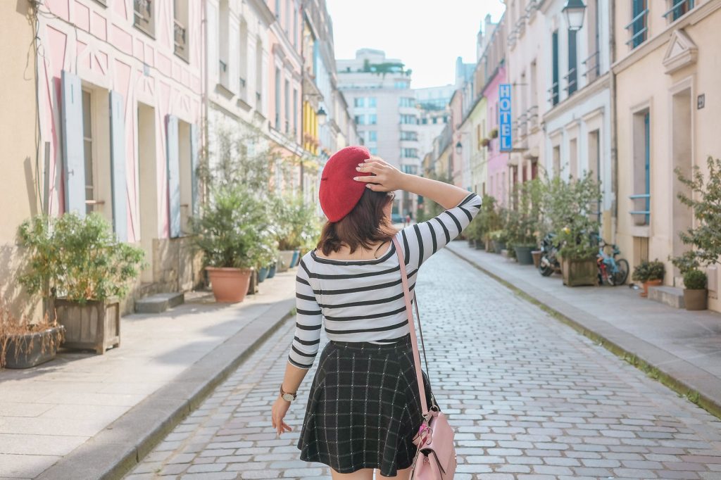 A girl in the middle of a cobbledstone road, admiring the pastel-colored buildings. The girl is wearing a red beret and striped top.